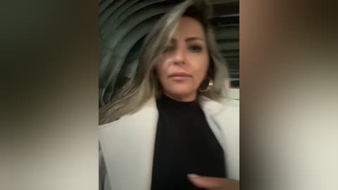 Media: A video of a woman with long, wavy, light brown hair, wearing a white blazer over a black turtleneck. She has a serious expression, with the background showing a blurred, modern interior.