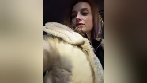 Media: Video of a woman with fair skin and light brown hair, wearing a beige puffy jacket, taken from a low angle. Background is dark and blurry, focusing on her face and upper body.