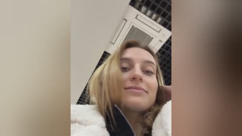 Media: Video of a young Caucasian woman with shoulder-length blonde hair, wearing a white fur-trimmed jacket, standing in a bathroom with tiled walls and an air conditioning unit above her.