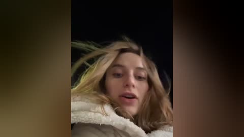 Media: Video of a young woman with long blonde hair, wearing a beige jacket, and a white, fluffy scarf, blowing in the wind, against a dark background.