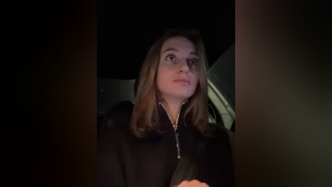 Media: Video of a Caucasian woman with shoulder-length, light brown hair, wearing a black jacket, looking contemplatively out of a car window. The background is dark, with the car interior faintly visible.