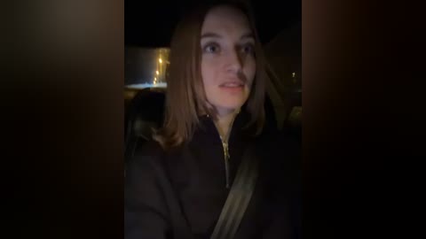 Media: Video of a young woman with straight brown hair, wearing a dark coat and a seatbelt, seated in a dimly lit car at night. The background is blurred and dark.