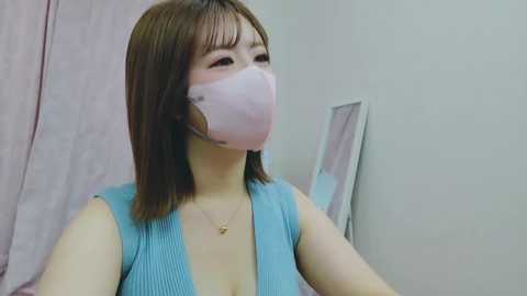 Media: Video of an East Asian woman with shoulder-length brown hair, wearing a blue sleeveless top, pink face mask, and a gold necklace. She is standing indoors against a pale wall, with a pink curtain and a white-framed mirror in the background.