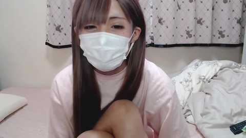 Media: Video of an East Asian woman with long dark hair, wearing a pink long-sleeve top, white surgical mask, and earbuds, sitting on a bed with white sheets, in a bedroom with floral-patterned curtains.