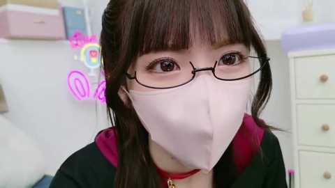 Media: A video of an East Asian woman with long brown hair, wearing glasses and a pink face mask, sitting in a brightly lit room with pastel-colored furniture and a colorful neon sign.