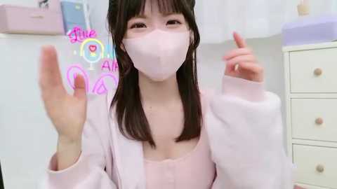 Media: Video of a young Asian woman with straight black hair and fair skin, wearing a pink face mask, long-sleeved top, and pink cardigan, smiling, in a light-colored room with pastel furniture.