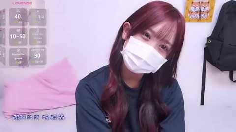 Media: Video of a young woman with long, wavy maroon hair and a white face mask, wearing a dark shirt, in a white room with a pink pillow, a clock, and a black bag.