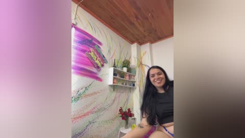 Media: Video of a cheerful young woman with long black hair, wearing a black crop top, smiling in a brightly lit room with colorful abstract wall art and a wooden ceiling.