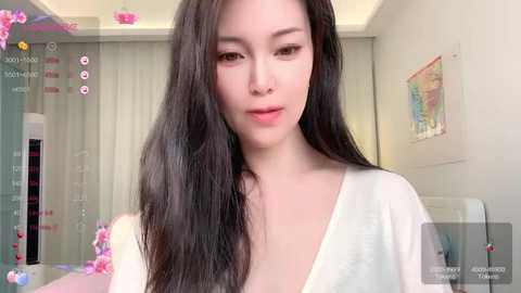 Media: Video of a young Asian woman with long black hair, fair skin, and a white top, smiling gently. Background features a white room with a window, colorful artwork, and floral decorations.