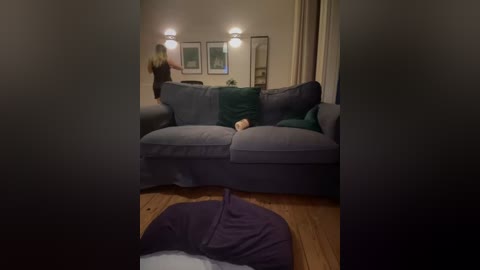 Media: Video of a cozy living room with a grey sectional sofa, green throw pillows, beige curtains, wooden floor, and a purple mat in the foreground.