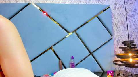 Media: Video of a modern, geometric headboard with light blue, diamond-shaped panels and a red light in the center. The background features a textured, light-colored wallpaper. A purple bottle and a candle are visible on a bedside table.