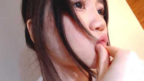 Media: A close-up video of an East Asian woman with long black hair, light skin, and dark eyes, biting her finger. The background is plain, possibly indoor with light wooden flooring.