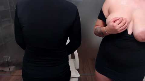 Media: Video of a person with fair skin, wearing a black long-sleeve shirt and a black dress, revealing their large breasts. The background features a tiled wall and wooden floor.