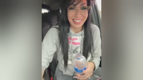 Media: Video of a smiling woman with long black hair, wearing glasses and a gray sweatshirt with \"I'm not a morning person\" in red text, holding a water bottle. She's sitting in a car with dark upholstery.