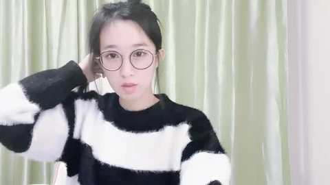 Media: Video of a young Asian woman with fair skin, dark hair, and round glasses, wearing a black and white striped sweater, standing in front of green, flowing curtains.