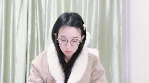 Media: Video of a young Asian woman with straight black hair, wearing glasses, a beige fur-trimmed coat, and light green curtains in the background.