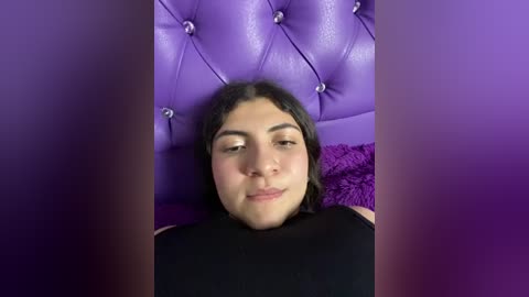 Media: Video of a young woman with medium brown skin and straight black hair, wearing a black sleeveless top, lying on a plush, tufted, deep purple velvet headboard with a textured, dark purple pillow.