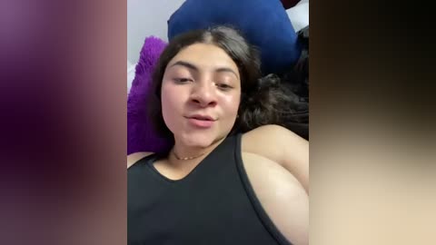 Media: Video of a young woman with medium skin tone, long dark hair, and a round face, lying on a bed, wearing a black tank top. She has a slight smile and her eyes are closed. Purple and blue pillows are visible in the background.