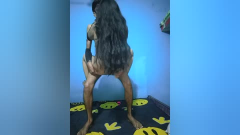 Media: Video of a person with long, dark hair and tan skin, wearing a tan bodysuit, squatting on a black yoga mat with yellow smiley faces.