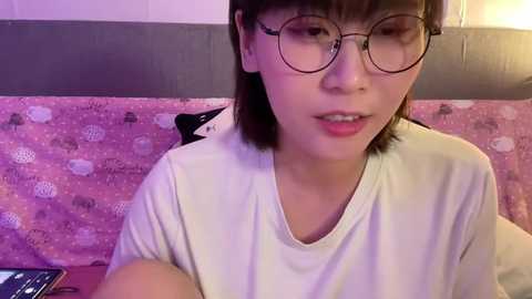 Media: Video of an Asian woman with short brown hair, wearing glasses and a white t-shirt, sitting in a pink and white polka-dotted chair.