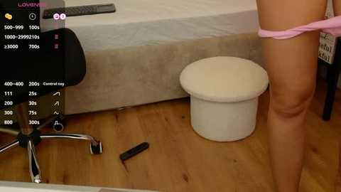 Media: A video showing a woman in pink panties, standing beside a white toilet with a black remote control on the wooden floor, in a modern bedroom with a bed and chair.