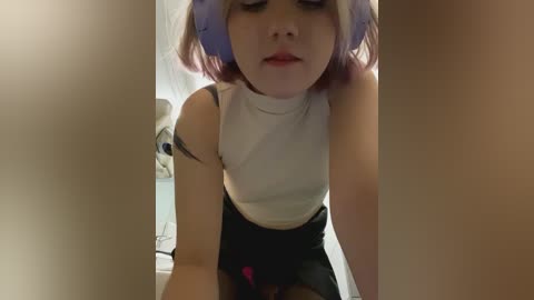 Media: Video of a young woman with light skin, pink hair, and purple headband, wearing a white tank top and black skirt, leaning forward, with a blurry background.