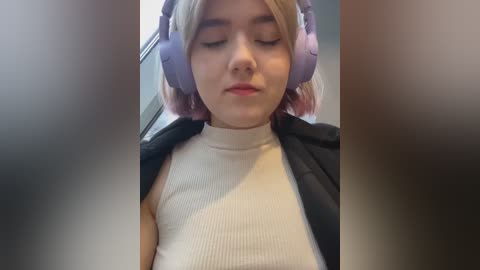 Media: Video of a young woman with fair skin, blonde hair, wearing large purple headphones, a beige ribbed tank top, and a black jacket, with a blurred background.