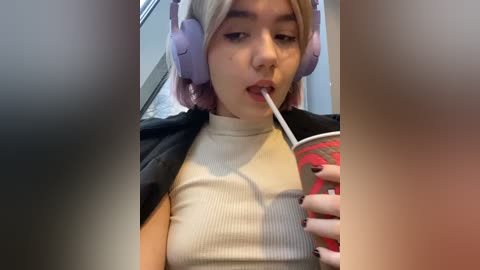 Media: Video of a young woman with light skin and short, dyed pink hair, wearing oversized lavender headphones and a ribbed white turtleneck, drinking from a red and white straw.