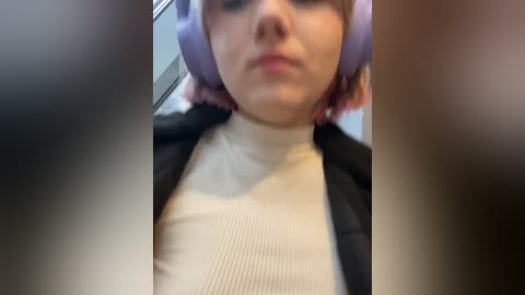 Media: Video of a woman with pale skin, short pink hair, wearing purple headphones, a black jacket, and a beige ribbed turtleneck sweater, sitting in a car. The background is blurry.