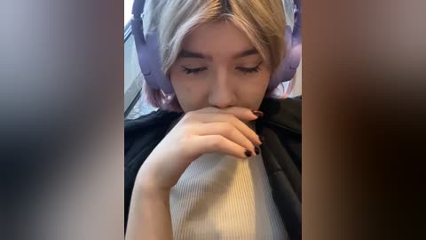 Media: Video of a young woman with light skin, blonde hair, and large purple headphones, covering her eyes as she holds her finger to her lips in a hush gesture. She wears a ribbed white top.