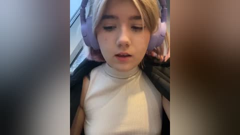 Media: Video of a young woman with blonde hair, wearing large purple headphones, a white ribbed tank top, and a black jacket, looking relaxed in a dimly lit car interior.