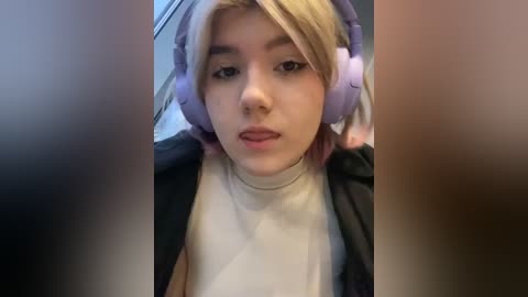 Media: Video of a young woman with fair skin and blonde hair, wearing large purple headphones and a black jacket over a white turtleneck. She has a neutral expression, with blurred, indistinct figures in the background.