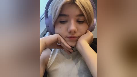 Media: A video of a young woman with fair skin and blonde hair, wearing large purple headphones and a sleeveless white top, resting her chin on her hands, with blurred faces in the foreground.