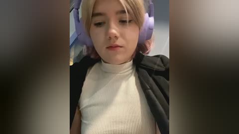 Media: Video of a young woman with fair skin and light brown hair, wearing a white ribbed turtleneck and black jacket, seated with headphones on, indoors.