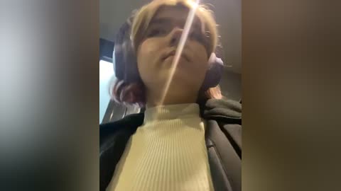 Media: A video of a young woman with shoulder-length blonde hair wearing large headphones, a black jacket, and a white turtleneck sweater, taken from a low angle, indoors, with a blurred background.