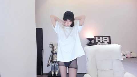 Media: Video of a slender, pale-skinned East Asian woman in a white T-shirt, black shorts, and black cap, posing in a minimalist room with white walls, a lamp, and a white armchair.