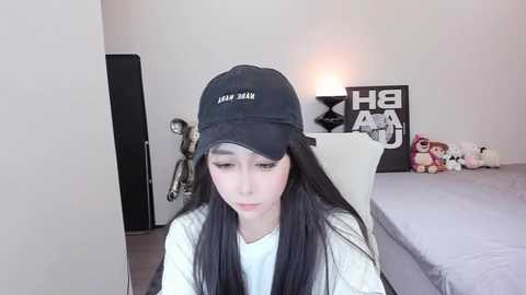 Media: Video of an Asian woman with long black hair, wearing a black cap and white top, sitting on a white chair in a minimalist bedroom with a bed, stuffed toys, and a black wall sign.