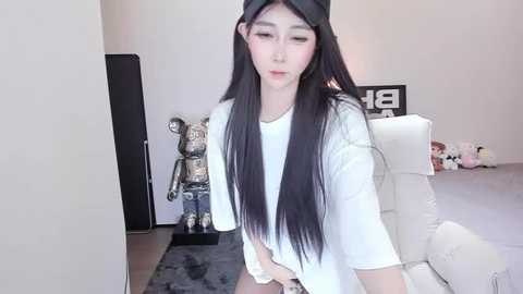 Media: Video of an East Asian woman with long, straight black hair, wearing a black beanie, white t-shirt, and shorts, sitting on a white couch in a modern, minimalist room with plush toys and a \"18\" sign.