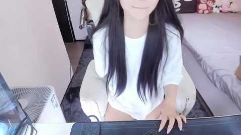 Media: Video of a young Asian woman with long black hair, wearing a white oversized sweater, sitting on a bed in a modern bedroom.