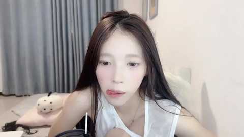 Media: A video of an East Asian woman with long black hair, wearing a white tank top, lying on a bed. The room has grey curtains, a plush toy, and a framed picture.