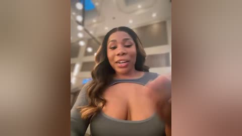 Media: Video of a smiling Black woman with long, wavy brown hair, wearing a gray top that exposes her ample cleavage, in a modern indoor setting with soft lighting and blurred background.