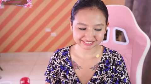 Media: Video of a smiling young woman with medium skin tone and straight black hair tied back. She wears a black floral blouse with blue accents, revealing a tattoo on her chest. Background features a pink gaming chair and a striped orange and yellow wall.