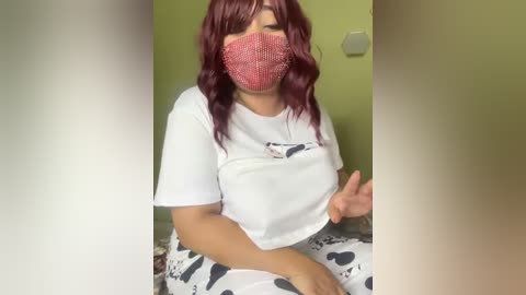 Media: Video of a woman with medium skin tone, long wavy maroon hair, wearing a white T-shirt and patterned pajama pants, and a pink face mask, sitting indoors.