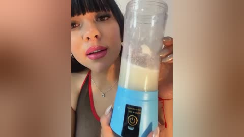 Media: A video of a young woman with medium skin tone and straight black hair, wearing a black top with red straps, holding a blue blender filled with a creamy substance.