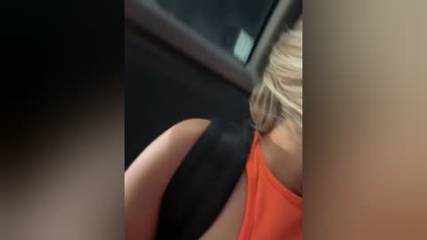 Media: Video: Close-up of a blonde woman's shoulder and arm in a bright orange top, seated in a car. The background is dark and blurry, emphasizing the subject's light-colored skin and attire.