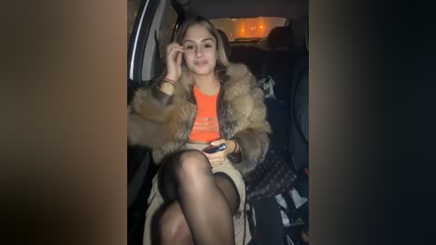 Media: Video of a young woman with light brown hair, wearing a fur coat, orange top, and sheer black stockings, sitting in a dimly lit car at night.