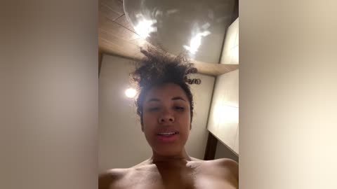 Media: A video of a topless Black woman with a curly afro, smiling, standing in a dimly lit room with wooden beams and a large, bright lamp.