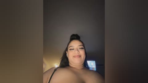 Media: A video of a smiling, plus-sized woman with medium brown skin and long black hair in a top knot, wearing a black bra, against a dark background.