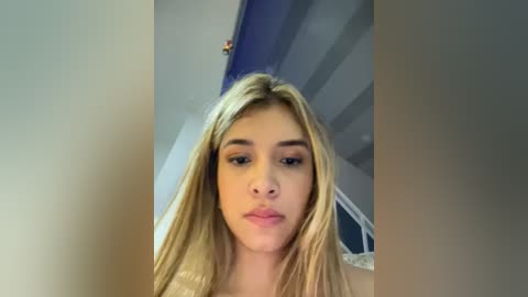 Media: A close-up video of a young Caucasian woman with long, straight blonde hair, fair skin, and light makeup, standing indoors in a dimly lit room with blue and white striped wallpaper.