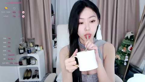 Media: Video of an Asian woman with long black hair, wearing a gray sweater, holding a white cup, standing in a cozy, lit room with a Christmas tree and a shelf filled with cosmetics.